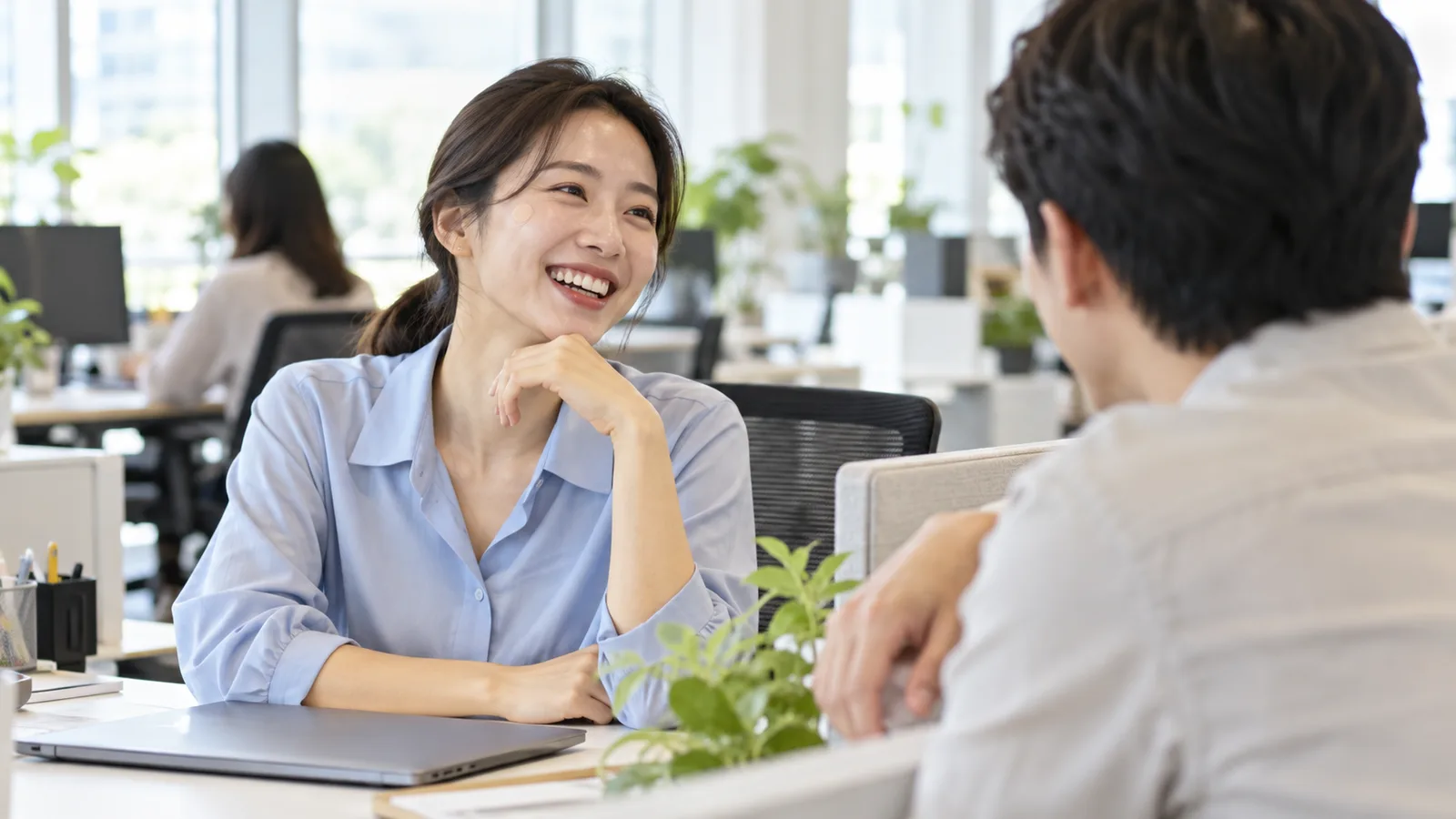 광대 옆에 작은 둥근 살색 밴드를 붙인 여성이 사무실 책상에서 동료와 웃으며 이야기하는 모습