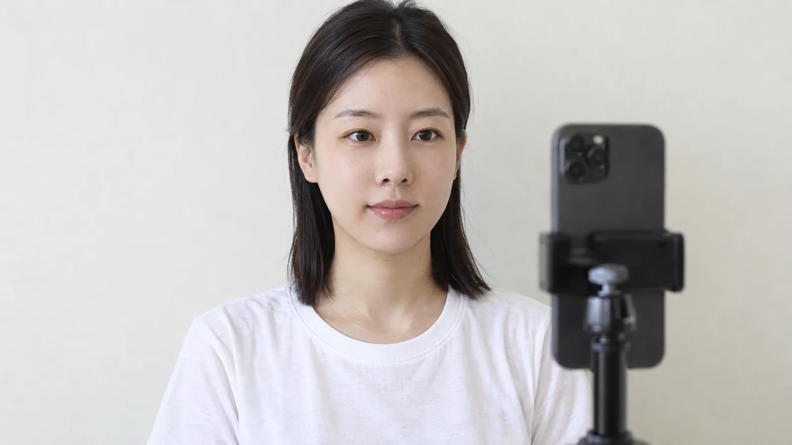 밝은 무지 벽 앞에서 성인 여성이 힘을 뺀 표정으로 정면을 바라보고, 옆에는 눈높이 거치대에 세운 휴대전화의 뒷면이 보이는 상반신 사진. 휴대전화 화면은 보이지 않는다.