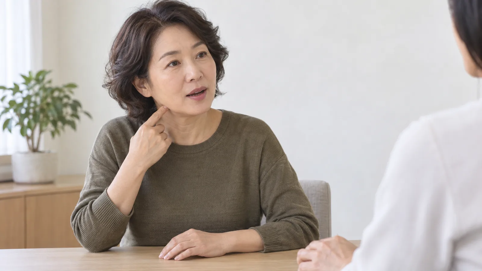 밝은 상담실 책상 앞에 앉은 중년 여성이 말하면서 검지로 턱선과 목 위쪽을 가리키고 맞은편 상담자는 어깨와 손만 일부 보인다.