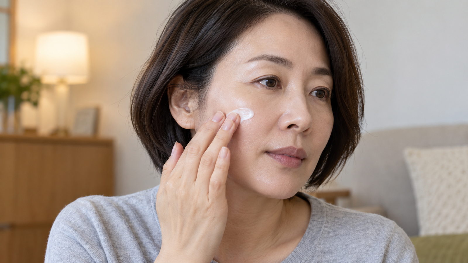 회색 옷을 입은 여성이 손끝에 묻힌 흰 크림을 볼에 바르는 모습